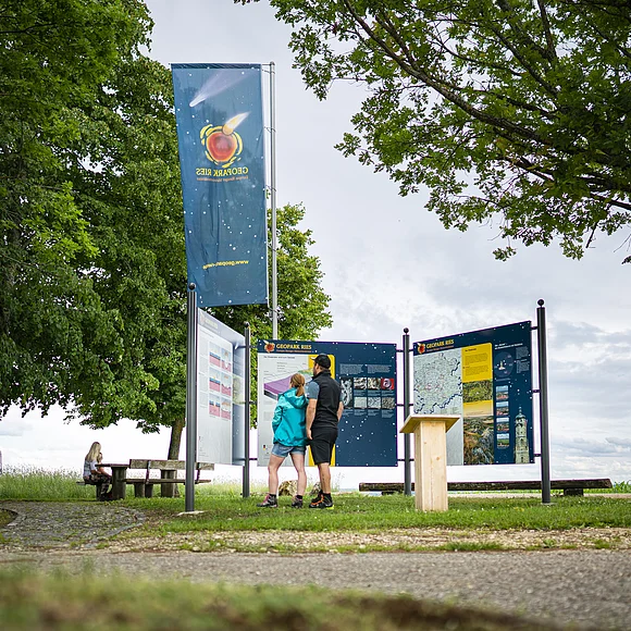 Zwei Personen betrachten Informationstafeln im Geopark Ries. Eine weitere Person sitzt auf einer Bank im Hintergrund.