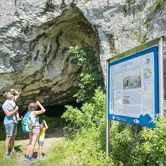 Eine Familie steht vor einer Höhle und betrachtet ein Informationsschild mit der Aufschrift "Ofnethöhlen".
