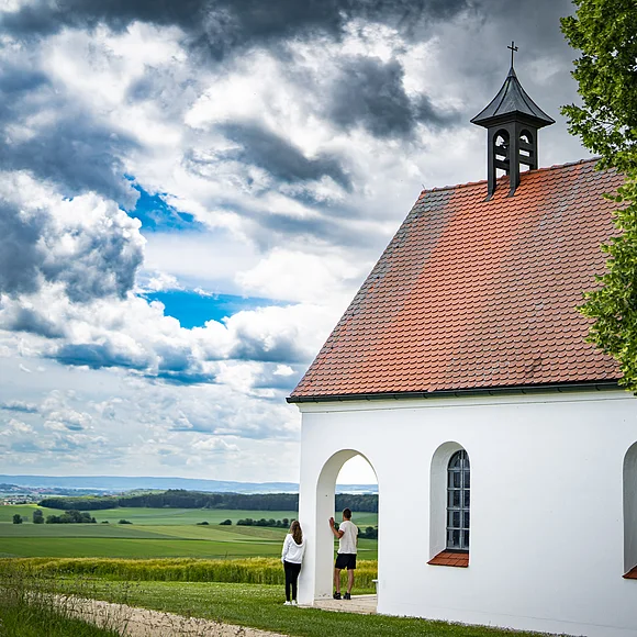 Kleine weiße Kapelle mit rotem Dach in ländlicher Umgebung, zwei Personen stehen davor.