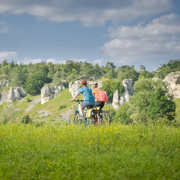 Zwei Personen fahren mit Fahrrädern auf einem grasbewachsenen Feld vor einer Felslandschaft.