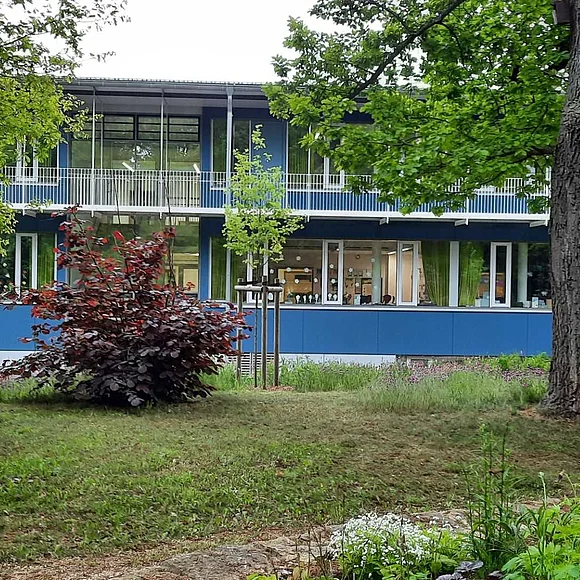 Zweistöckiges Gebäude mit blauer Fassade, umgeben von Bäumen und Sträuchern. Fenster im Erdgeschoss dekoriert.