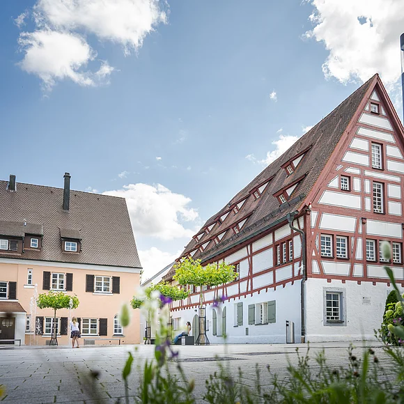 Fachwerkhaus mit rotem Giebel und weißen Wänden, daneben ein helles Gebäude, blauer Himmel im Hintergrund.