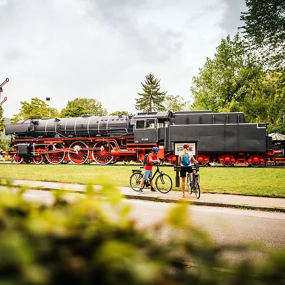 Dampflokomotive auf Gleis mit zwei Radfahrern davor, umgeben von Bäumen und Wiese bei bewölktem Himmel.