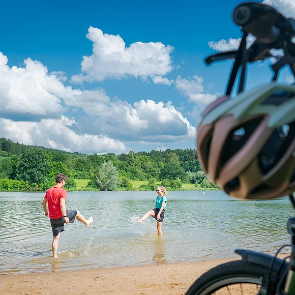Im Vordergrund stehen rechts zwei Fahrräder, dahinter liegt ein kurzer Strand an einem Seeufer. Im seichten Wasser stehen ein Mann und in eine Frau in kurzen Hosen und T-Shirts, die versuchen, sich gegenseitig nass zu spritzen. Am Ufer gegenüber liegen eine Wiese und Wald.