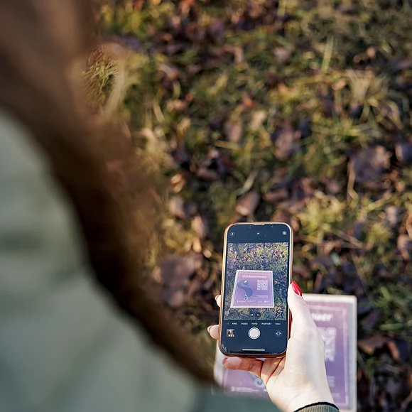 Person fotografiert mit Smartphone eine Tafel mit Text und QR-Code auf Grasboden im Freien.