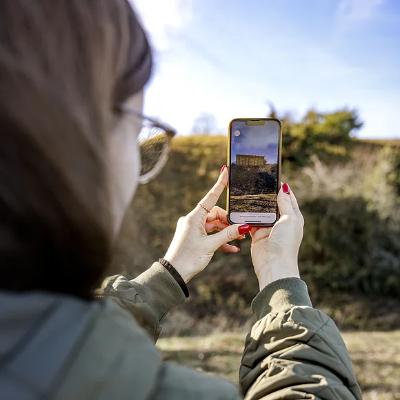 Frau mit Brille und rotem Nagellack hält Smartphone mit Bild einer historischen Pfostenschlitzmauer im Freien.