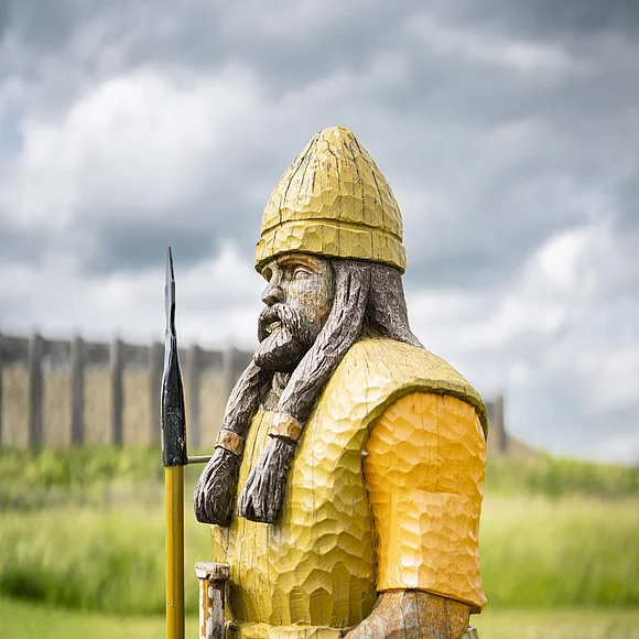 Auf einer Wiese steht die aus Holz geschnitzte und bemalte Figur eines bewaffneten keltischen Mannes. Im Hintergrund ist ein rekonstruiertes Bauwerk zu sehen.