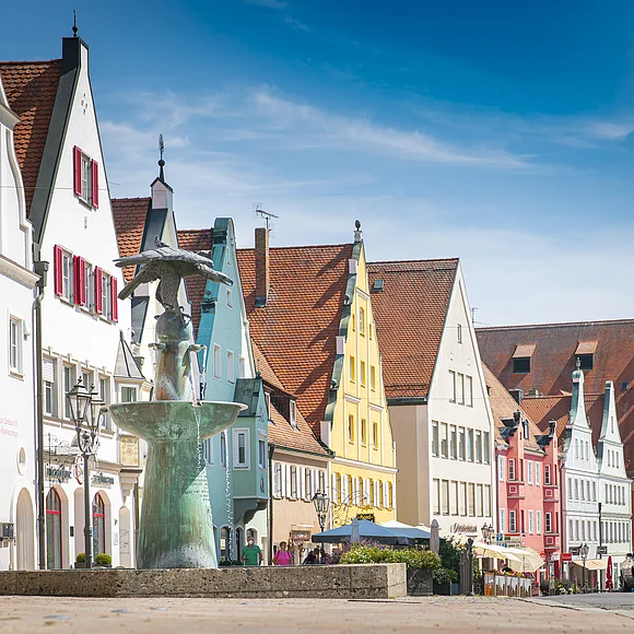 Straße mit bunten Giebelhäusern und einem Brunnen im Vordergrund. Klarer Himmel im Hintergrund.
