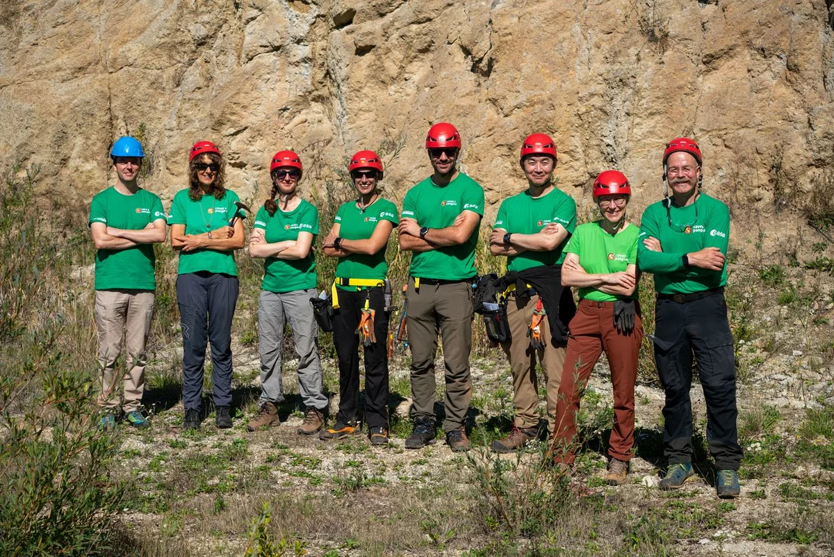 Teilnehmende und Lehrende des PANGAEA-Trainings Auf dem Bild sind acht Menschen mit Helmen und grünen T-Shirts zu sehen, die ihre Arme verschränkt haben. Sie stehen im Gras vor einem großen braunen Felsen.